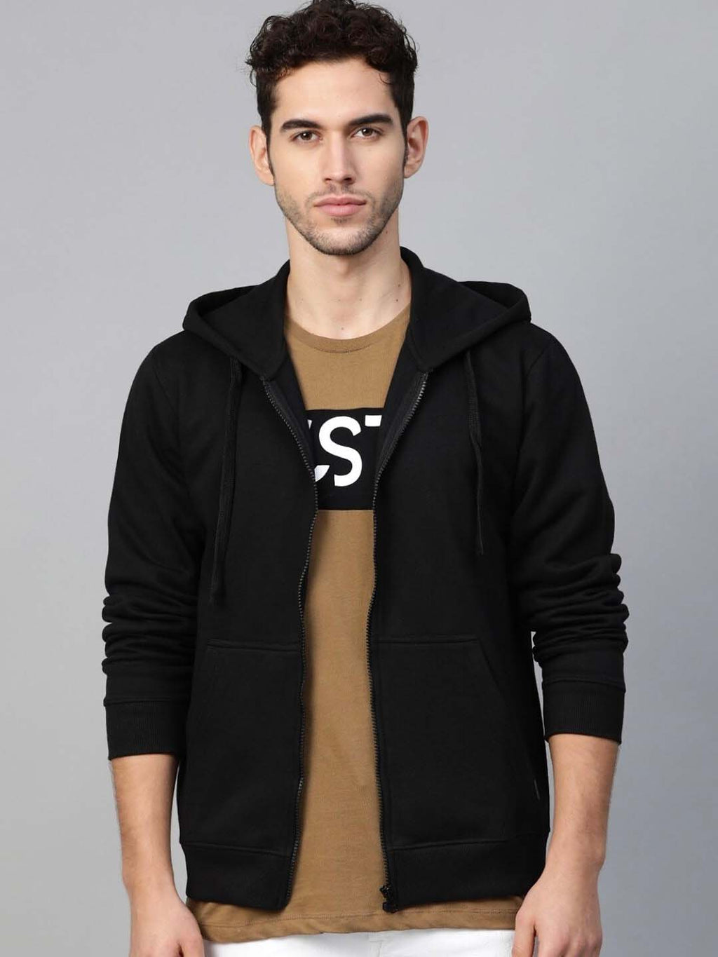 A man wearing a black zip-up hoodie over a brown graphic t-shirt, standing against a solid gray studio background.