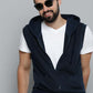 A man wearing a sleeveless navy blue zip-up hoodie vest over a white t-shirt and dark jeans, accessorized with black sunglasses, posed against a solid gray studio background.