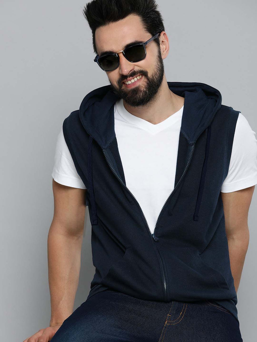 A man wearing a sleeveless navy blue zip-up hoodie vest over a white t-shirt and dark jeans, accessorized with black sunglasses, posed against a solid gray studio background.