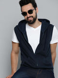 A man wearing a sleeveless navy blue zip-up hoodie vest over a white t-shirt and dark jeans, accessorized with black sunglasses, posed against a solid gray studio background.