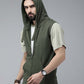 A man wearing an olive green sleeveless zip-up hoodie vest over a light beige t-shirt and black pants, photographed against a solid gray studio background.