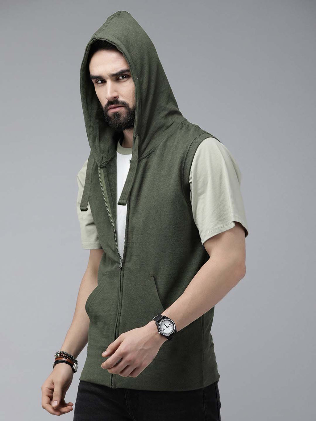 A man wearing an olive green sleeveless zip-up hoodie vest over a light beige t-shirt and black pants, photographed against a solid gray studio background.