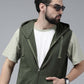 A man wearing a dark olive green zip-up hooded vest over a white t-shirt and light olive green sleeves, styled with sunglasses and a watch, against a solid gray studio background.