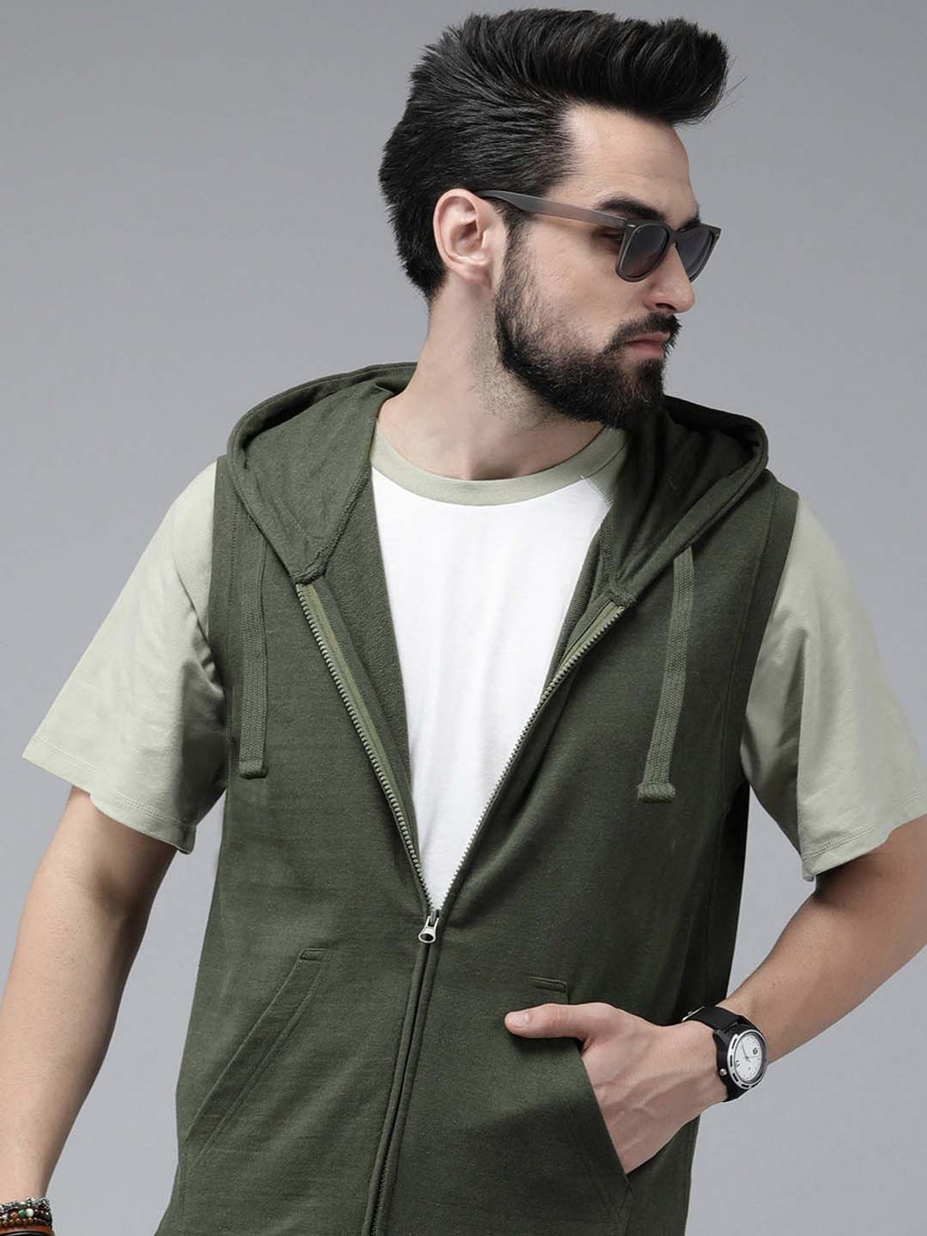 A man wearing a dark olive green zip-up hooded vest over a white t-shirt and light olive green sleeves, styled with sunglasses and a watch, against a solid gray studio background.