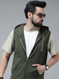 A man wearing a dark olive green zip-up hooded vest over a white t-shirt and light olive green sleeves, styled with sunglasses and a watch, against a solid gray studio background.