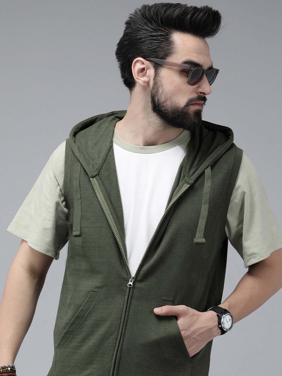 A man wearing a dark olive green zip-up hooded vest over a white t-shirt and light olive green sleeves, styled with sunglasses and a watch, against a solid gray studio background.