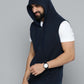 A man wearing a sleeveless navy blue zip-up hoodie vest over a white t-shirt, paired with dark jeans and a black watch, photographed against a solid gray studio background.