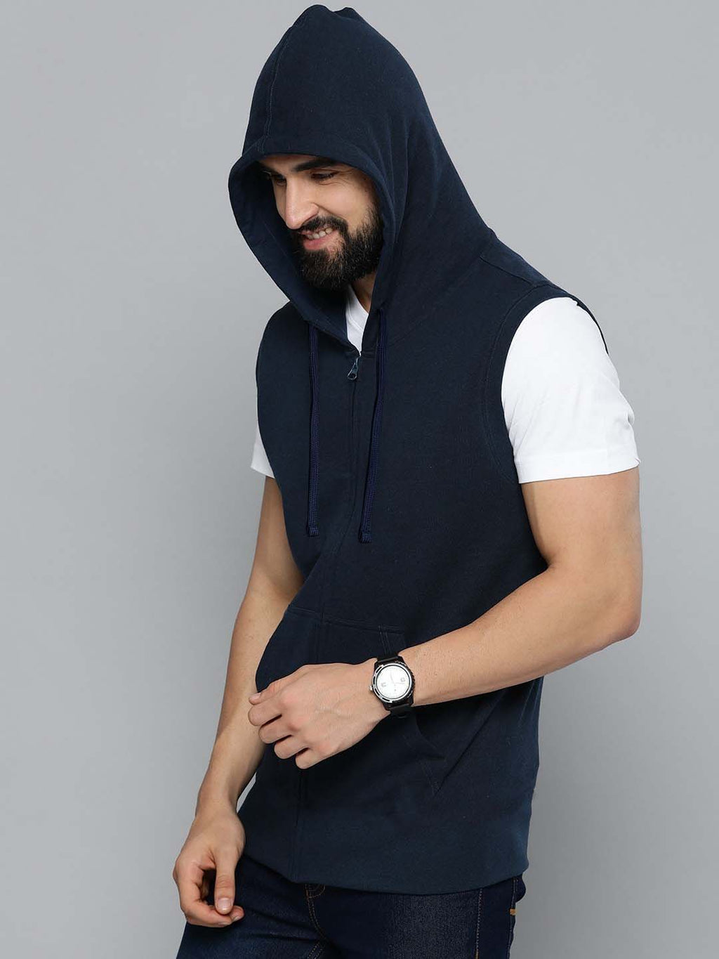A man wearing a sleeveless navy blue zip-up hoodie vest over a white t-shirt, paired with dark jeans and a black watch, photographed against a solid gray studio background.