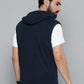 A man shown from the back wearing a sleeveless navy blue hooded shirt layered over a white short-sleeve t-shirt, set against a plain gray studio background.