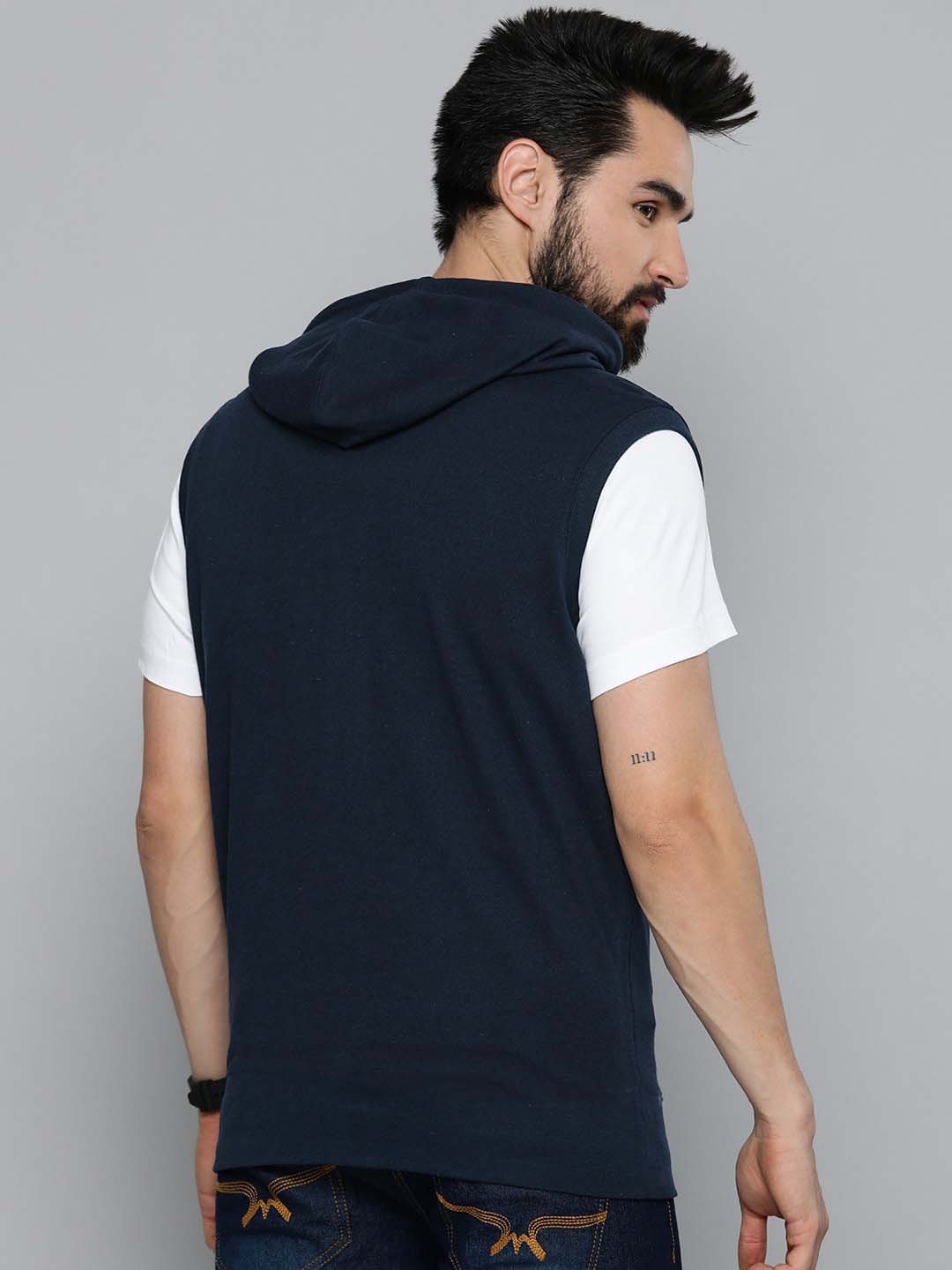 A man shown from the back wearing a sleeveless navy blue hooded shirt layered over a white short-sleeve t-shirt, set against a plain gray studio background.
