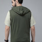 A man shown from the back wearing a sleeveless olive green hooded shirt layered over a light beige t-shirt, set against a plain gray studio background.
