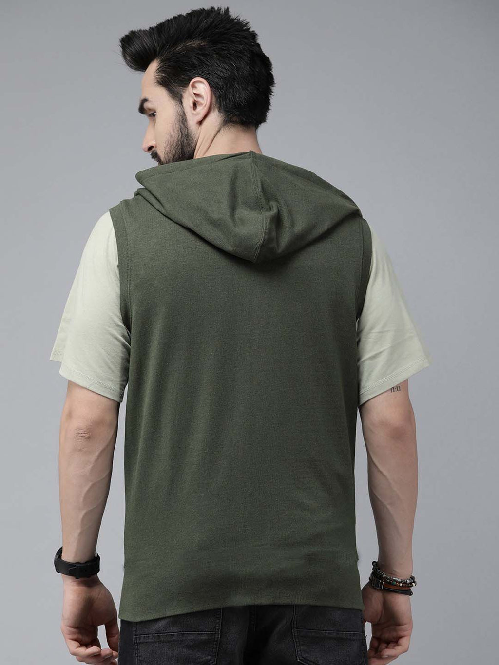 A man shown from the back wearing a sleeveless olive green hooded shirt layered over a light beige t-shirt, set against a plain gray studio background.