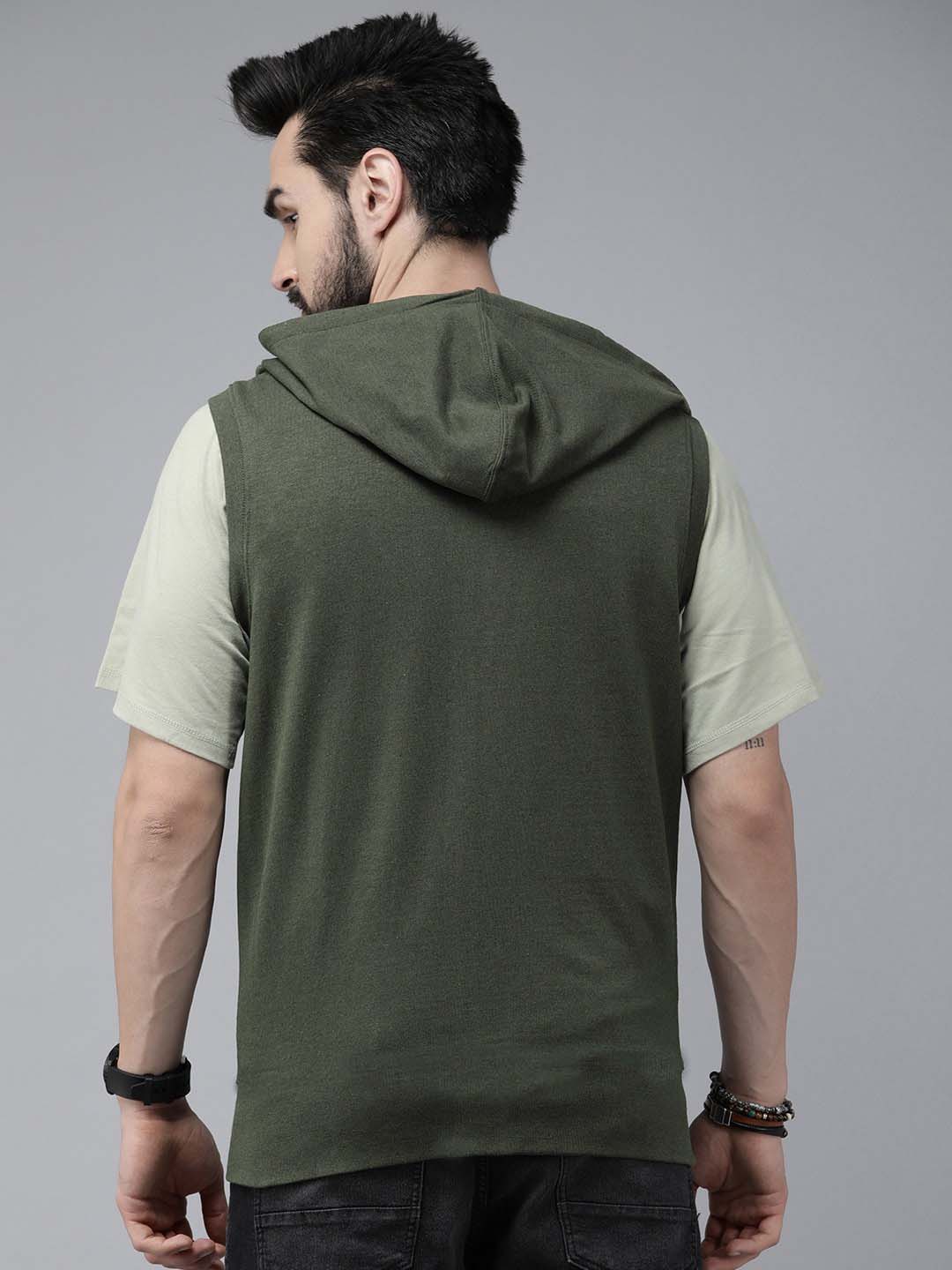 A man shown from the back wearing a sleeveless olive green hooded shirt layered over a light beige t-shirt, set against a plain gray studio background.
