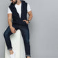 A man wearing a navy blue zip-up hoodie vest over a white t-shirt, dark blue jeans, and white sneakers, sitting on a white block against a gray studio background.