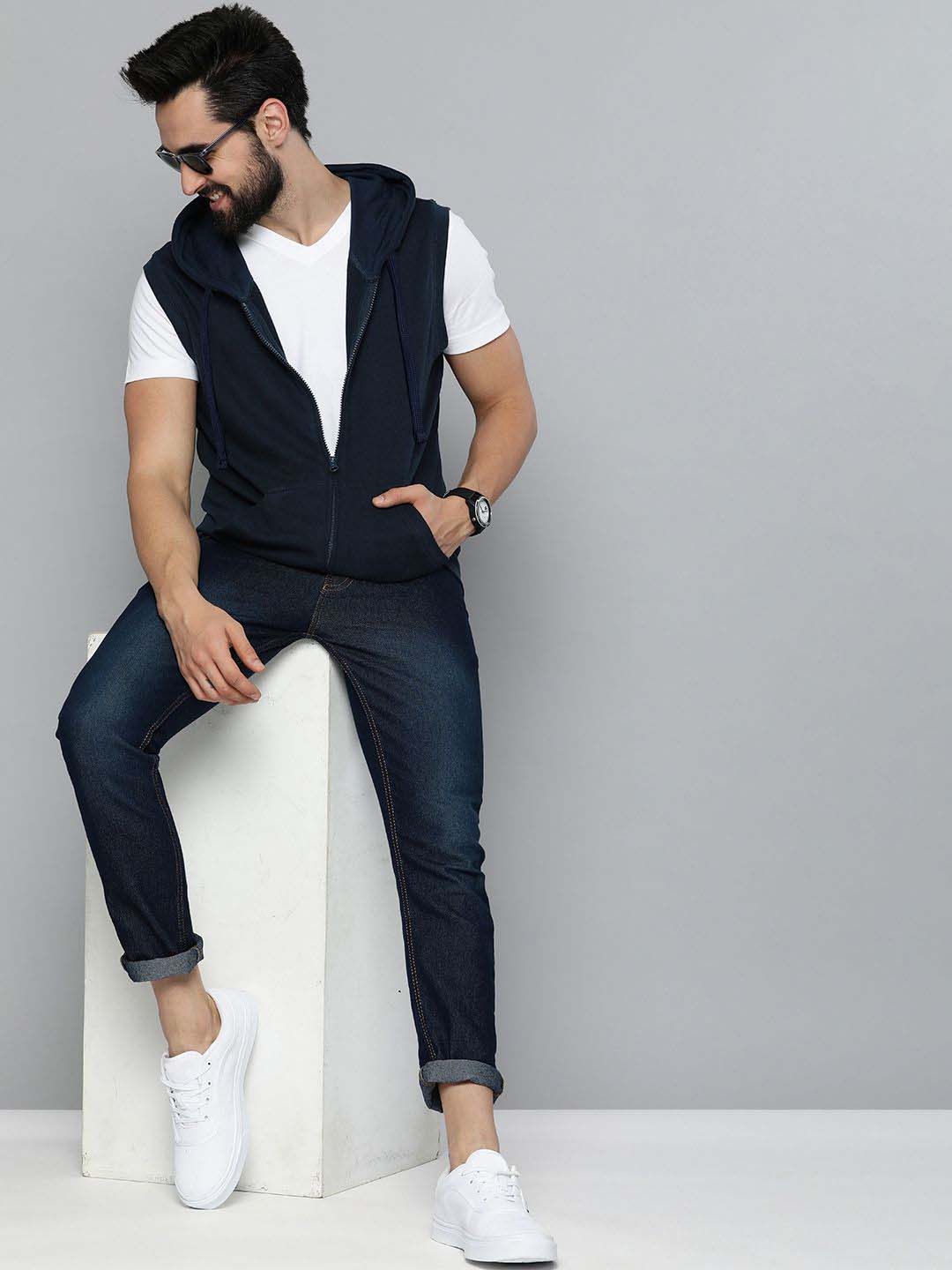 A man wearing a navy blue zip-up hoodie vest over a white t-shirt, dark blue jeans, and white sneakers, sitting on a white block against a gray studio background.