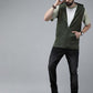 A man modeling a sleeveless olive green zip-up hoodie vest over a light green t-shirt, black jeans, and black boots, standing against a solid gray studio background.