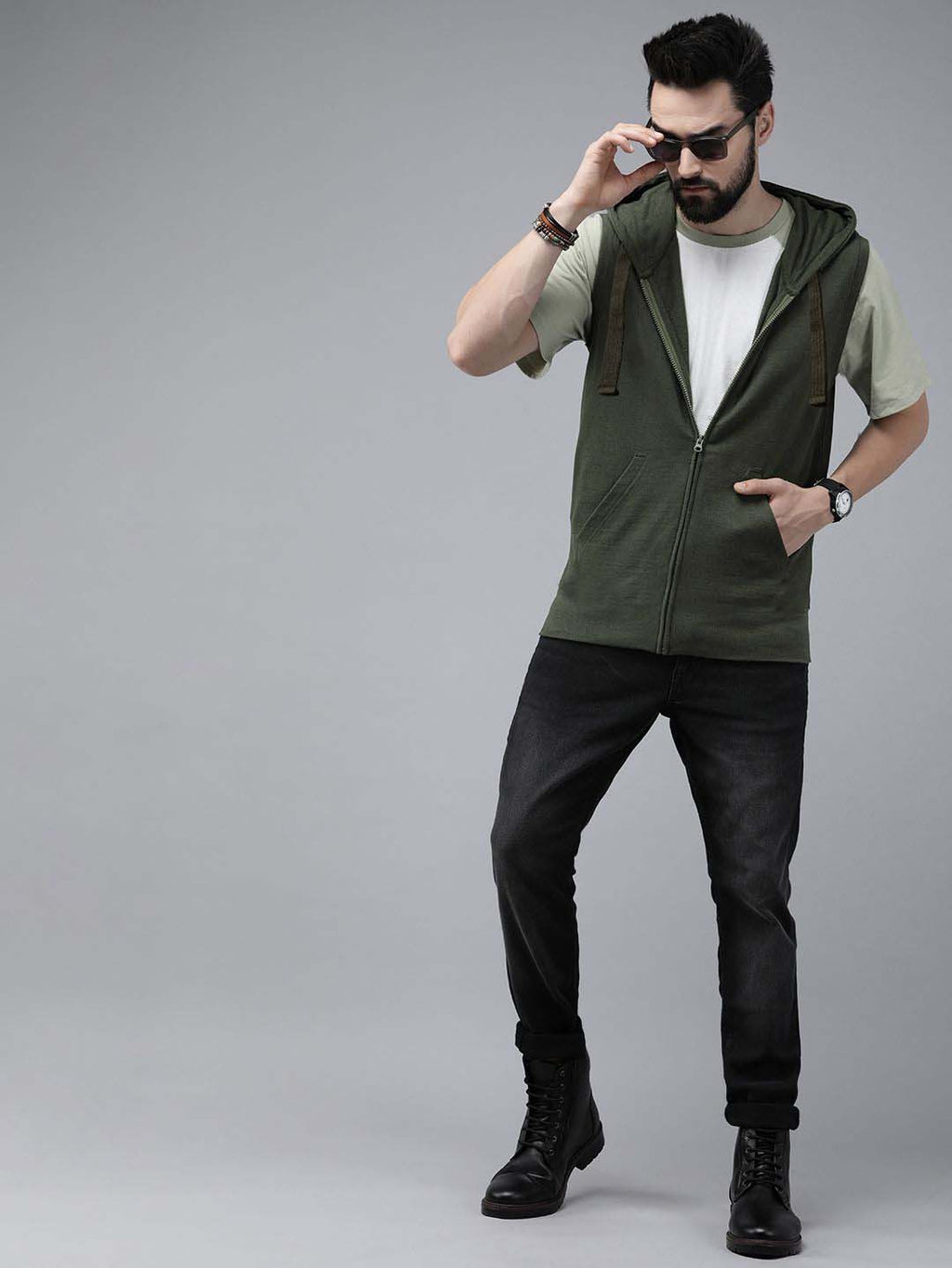 A man modeling a sleeveless olive green zip-up hoodie vest over a light green t-shirt, black jeans, and black boots, standing against a solid gray studio background.