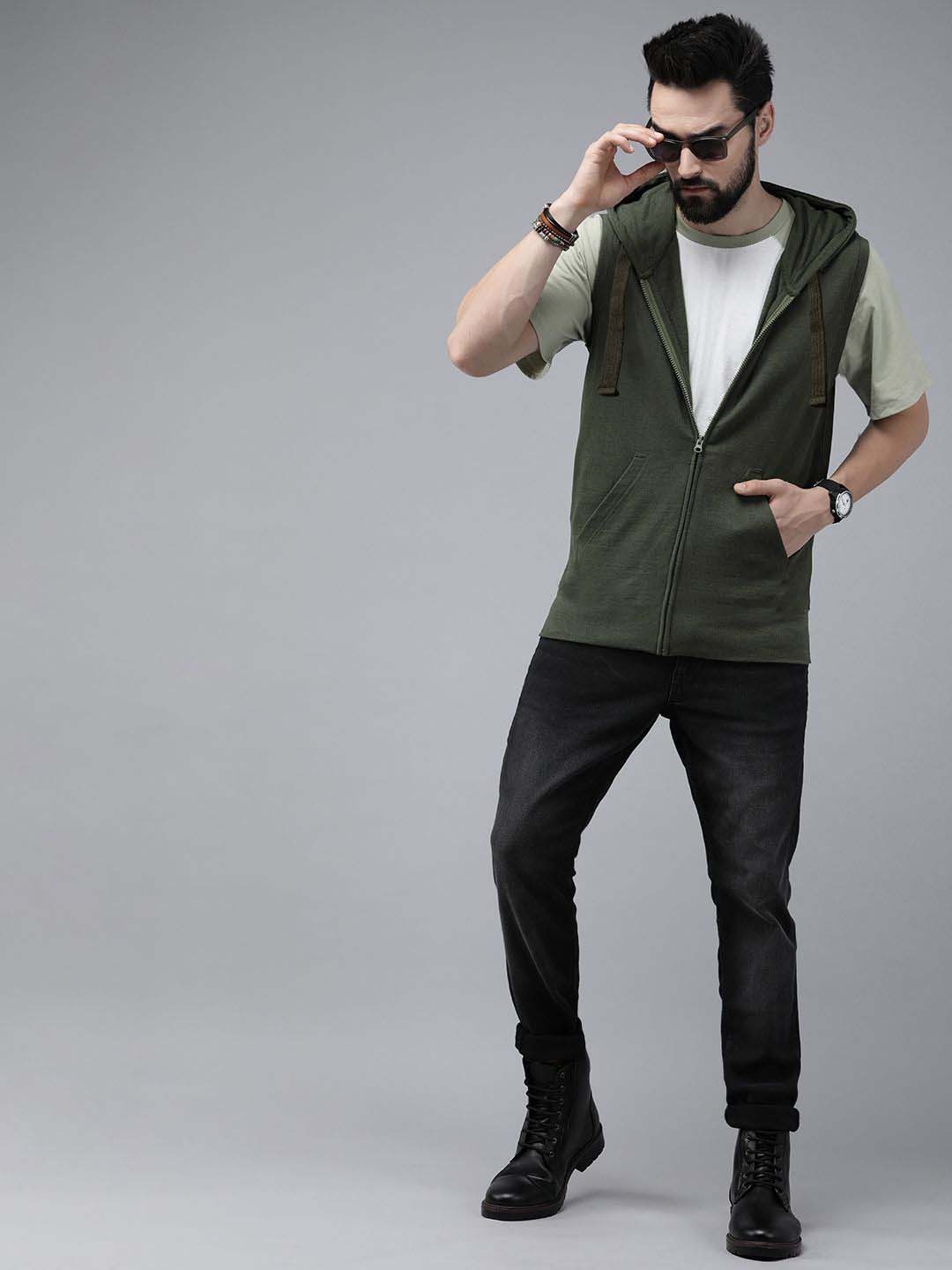 A man modeling a sleeveless olive green zip-up hoodie vest over a light green t-shirt, black jeans, and black boots, standing against a solid gray studio background.